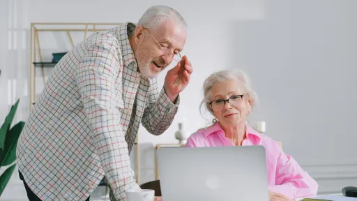 A man and woman are looking at a laptop. The man is standing next to the seated woman. He is smiling and holding his glasses to his head. The woman is seated at a desk with a laptop, notebook and a tea cup.