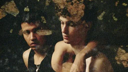 A photograph of two male figures sitting side by side against a black background. One has his arm over the shoulder of the other and is looking at him. The other is looking downwards and is holding a pale pink rose. Patches of semi-transparent brown botanical pattern cover the photograph. 
