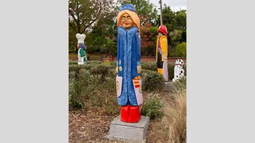 A carved wooden figure of a glamorous woman with a long blue coat. She has blond hair, bright red boots and carries two shopping bags.