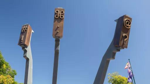 : Three arms that mimic the arms of a typewriter are reaching towards the sky. They carry symbols like an at sign and an exclamation mark’