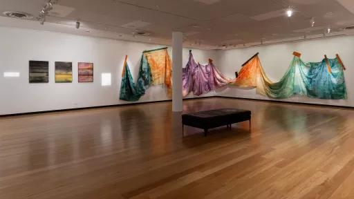 A gallery installation with dyed fabric draped and suspended, with small abstract paintings on the opposite wall.