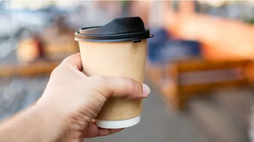 Person's hand holds disposable paper coffee cup with plastic lid