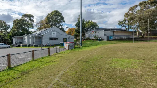The edge of a sportsground oval with line markings and a short wire fence. 2 buildings are next to the oval. 