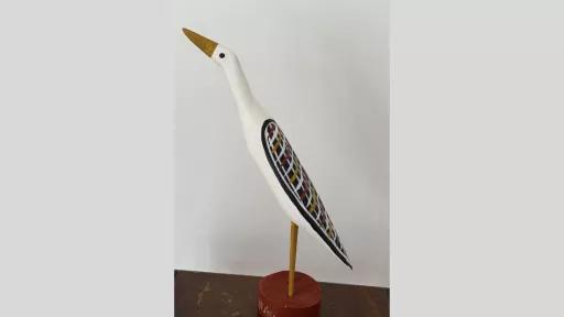 Image of a carved wooden bird painted white with a yellow beak and black eye, mounted on a red circular base. Its wings feature a geometric cross-hatch design in red, white, yellow, and black.
