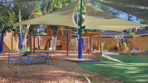 Shaded outdoor play area with climbing equipment, swings, and artificial turf, surrounded by trees and a wooden fence.equipment