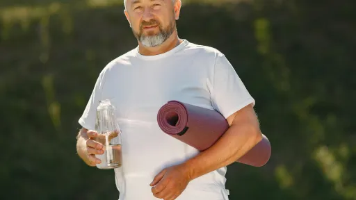 A man is wearing a white tshirt holding a drink bottle in one hand and a yoga mat tucked under the other arm.
