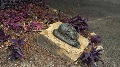 A small bronze sculpture in a garden bed, featuring the side of a young girl’s face and her arm. The bronze is attached to a sandstone base