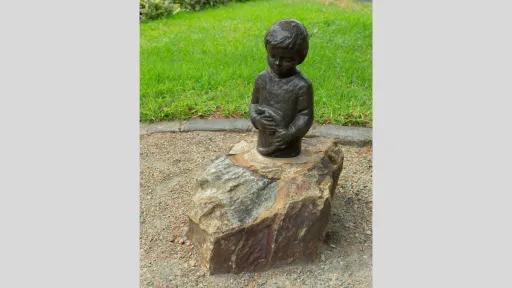 A bronze bust of a child holding a lizard. Thue bust is attached to a large stone base in a garden