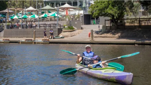 Woman and child wearing life vests in kayak on river outside building with café umbrellas.