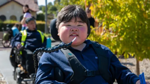Three people in wheelchairs on path in playground, with boy smiling at front and house and trees in background.