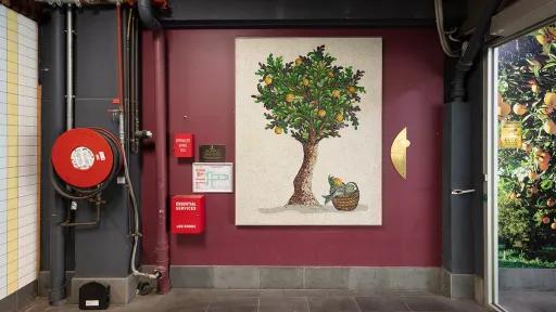 text: A photograph of a mosaic hanging on a purple wall. The mosaic features a tree. There are utilities surround the mosaic including a fire hose.