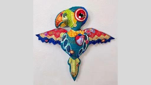 A colourful, hand-stitched textile sculpture of a stylised bird with wings outstretched. The bird is constructed from multicoloured fabric with floral and geometric patterns, embroidery, and applique. It is mounted on a white wall.