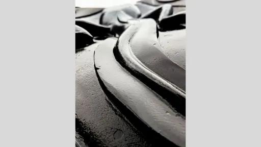 An extreme close-up of a dark, glossy surface with curved, raised patterns. The surface appears smooth and reflective, with highlights that suggest a wet or coated texture.