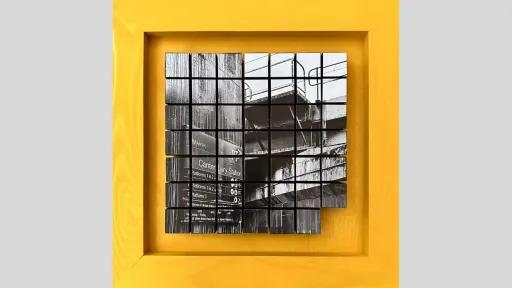 A three-dimensional artwork framed in yellow. Inside the frame is a grid of black and white photographic tiles that form an image of Canterbury Station. The photograph shows a concrete railway overpass and signage, with the grid creating a fragmented effect.