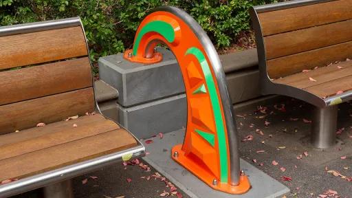 Orange and green public sculpture in a rainbow shape between 2 park benches