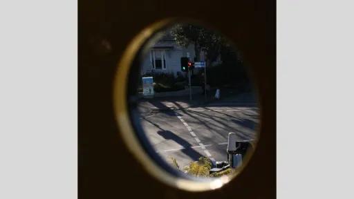 An urban street scene viewed through a small circular opening. The image frames a green traffic light, a pedestrian, and the corner of a house. Shadows fall across the road.