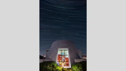 A modern white building with a large stained-glass window centered in the frame. The night sky above is captured in a long exposure, showing streaked star trails curving across the image.
