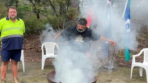 Smoking billowing out of a fire pit as an Aboriginal man conducts a smoking ceremony.