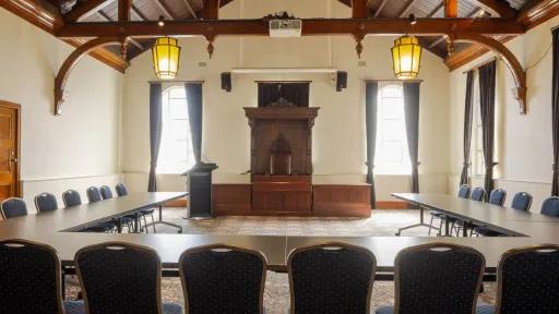 A conference room with long tables arranged in a U-shape, facing a raised platform with a wooden podium and antique-style furniture. The room has high, wood-beamed ceilings and arched details.