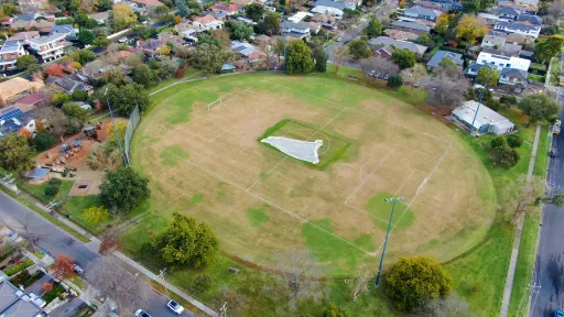 An aerial view of a oval sportsground. It is surrounded by a playground and houses.