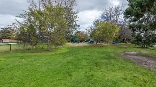 A grass area with. A sportsground fence, trees and a play area is in the background