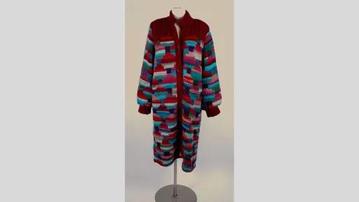 Photograph of a long-knitted coat dress displayed on a mannequin, featuring a button-down front and a high, ribbed collar. The garment has a red base with multicoloured block patterns in shades of teal, pink, purple, and grey. The cuffs are ribbed and gathered, and the coat has a loose, straight silhouette that extends to mid-calf length.