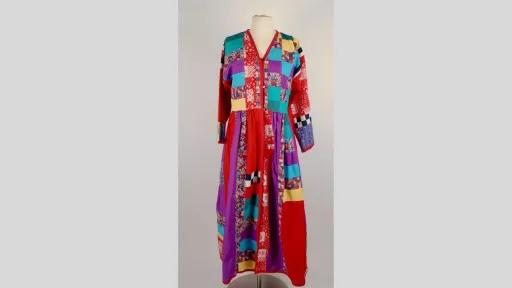 Photograph of a long-sleeved, ankle-length dressing gown displayed on a mannequin. The garment is constructed from a variety of colourful fabric patches, including florals, checks, polka dots, and solid colours like red, yellow, turquoise, purple, and black and white. The dress features a V-neckline, a buttoned bodice, and a gathered waist, creating a flared lower half.