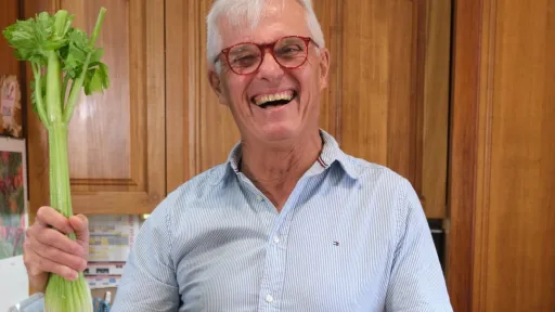 A middle-aged man in a wooden kitchen beams as he holds a stalk of celery