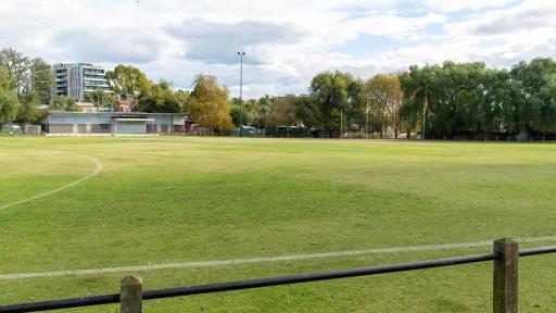 A playing field with a low fence. It is surrounded by trees. Footy goal posts and a small building are on the far side of the field