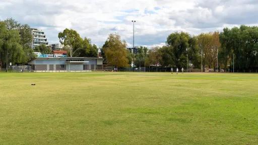 A green sports field with trees, goalposts and a buiding in the background.