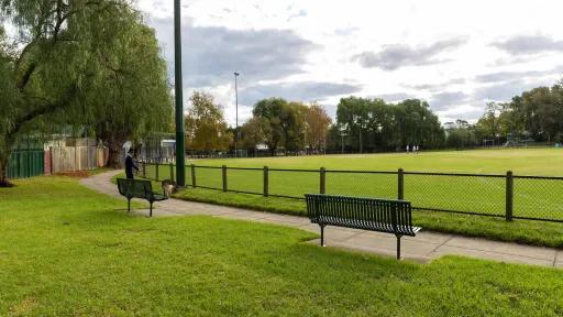 A park with benches and a grassy field. The park is fenced and it is surrounded by a walking path.