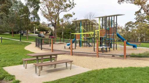 An outdoor playground with chain'link fencing featuring a climbing structurem and a slide. Outside the playground is a good picnic table.