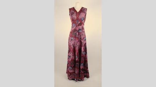 Photograph of a floor-length, sleeveless dress displayed on a mannequin. The fabric features an all-over pattern of large, floral design in shades of pink, purple, and black. The dress has a V-shaped neckline with overlapping panels at the bust and a fitted bodice that flares into a wide, flowing skirt.