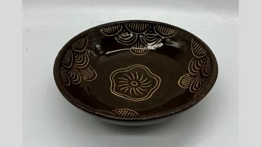 Photograph of a shallow, round ceramic dish with a dark brown glossy glaze. The interior surface is decorated with carved out floral and fan-like patterns in a lighter beige colour. 