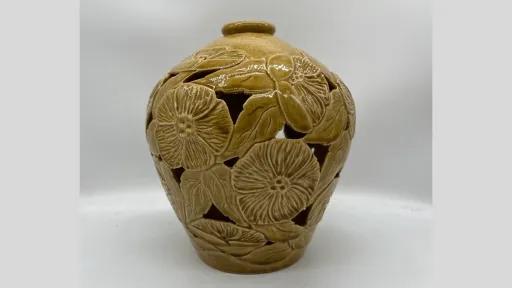 Photograph of a bulbous, light brown ceramic vase with a small opening at the top. The surface features carved floral and leaf motifs, with cut-out sections between them that create a lace-like design. The glaze is smooth and glossy.