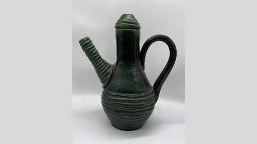 Photograph of a ceramic vessel with a deep green glaze, featuring a tall, narrow neck and a pointed lid. The spout is short and angled, with a ridged, spiral design. The body is rounded and decorated with evenly spaced horizontal ridges, and a single curved handle extends from the upper neck to the midsection.