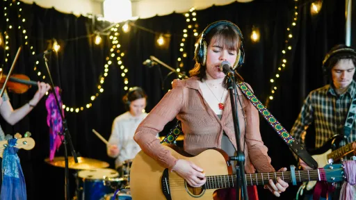 A 4-piece band play musical instruments. Decorative fairylights can be seen in the background. 