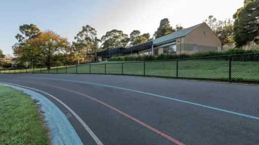 A close up of a cycling track with yellow, red and blue markings surrounded bu a short wire fence. There is a building and trees in the background. 
