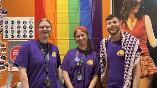 Three youn people in purple t-shirts standing in front of a pride flag hung on the wall behind them