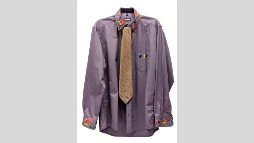 Photograph of a purple collared shirt. Collar, cuff of sleeves, and pocket are painted with colourful graphic shapes in red, yellow, blue, green and white. On top is a golden tie around the collar, painted with blue diagonal lines. 