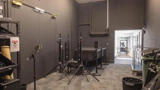 A windowless room that is painted dark grey and contains equipment like speaker stands, music stands, a table, and rolling carts. A hallway is visible through a doorway.