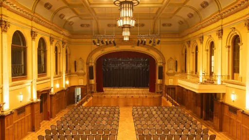The spacious auditorium's interior includes a stage. Rows of chairs are set up in the main floor. There is a balcony on the right side and a large stage with red curtains at the far end. The room has ornate architectural details and is lit with chandeliers.