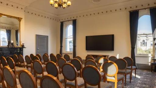 A meeting room featuring rows of wooden chairs with gray fabric seats, arranged in front of a large, wall-mounted television. Two windows flank the screen, draped with dark curtains. An ornate fireplace and mirror are visible to the left. The room has detailed ceiling molding and a chandelier.