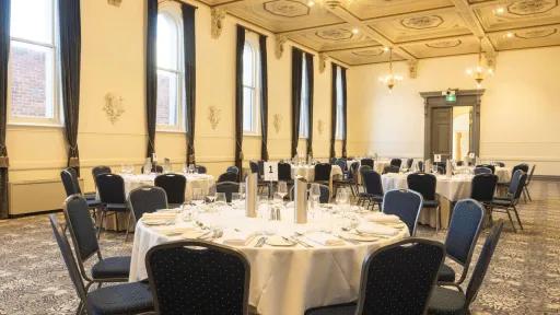 curtains adorn the walls. A spacious room is arranged for a banquet or large dinner. Round tables are adorned with white tablecloths and blue and silver chairs. Each place setting includes silverware and glasses. The ceiling features intricate, decorative molding, and chandeliers. The walls have tall windows, adorned with dark curtains. A doorway is visible in the background. The room is carpeted with a floral pattern in various shades of grey.
