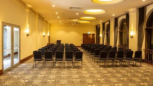 A spacious conference room with blue chairs arranged in rows facing a podium. The room features floral-patterned carpeting, pale yellow walls, and arched windows with dark drapes. A door with glass panes is on the left.