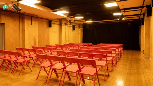 A small event space with rows of red folding chairs facing a black curtained stage. The walls are wood paneled and there are lighting fixtures and speakers mounted on black tracks on the ceiling. 