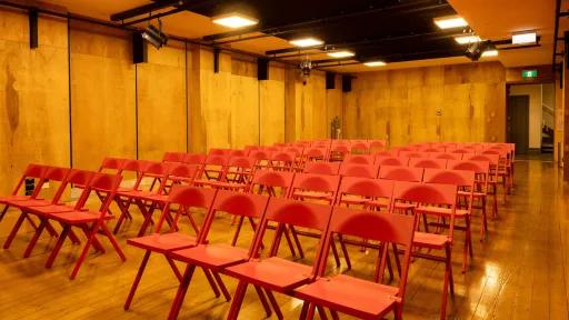 A room prepared for a presentation. Rows of red folding chairs are arranged on a wooden floor facing the back of the room, with wood paneling. The black ceiling features square lights and stage lights. A doorway with a green exit sign is situated on the right side of the room.