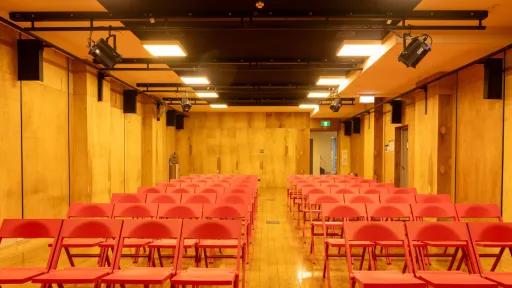 Red chairs are arranged in rows facing the front inside an event space. The walls are constructed of wooden panels and illuminated by square ceiling lights and stage lighting.
