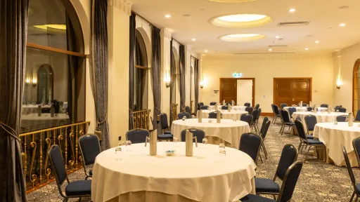 A conference room is set up with round tables covered in white cloths, each surrounded by chairs with blue patterned upholstery. There are large, arched windows with dark curtains along one side of the room. The ceiling features recessed lighting and a projector. The floor is covered with a patterned carpet.