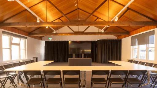 A meeting room with a large U-shaped table, black chairs, a wood-paneled ceiling, and a large monitor on a stand at the front.